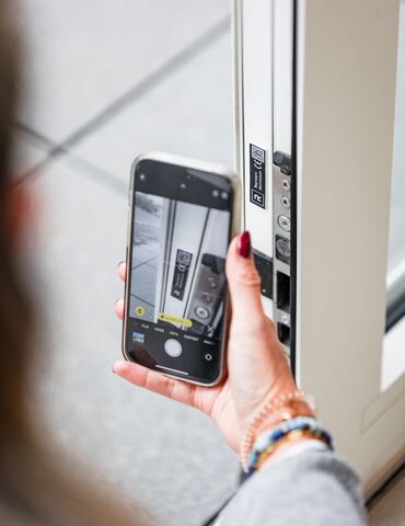 Kéz tartja az okostelefont a QR-kód közelében, fehér ablakprofilon.